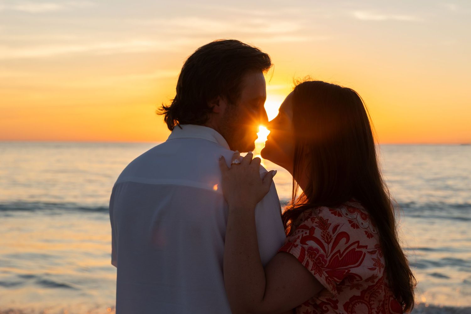 Silhouetted couple share intimate moment at beach during golden sunset over ocean horizon.