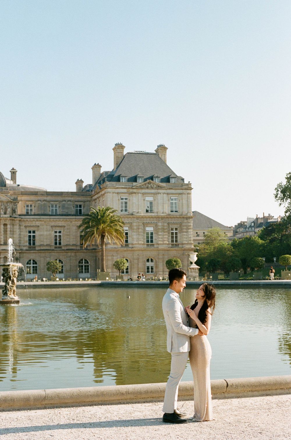 A couple embraces in front of the historic Luxembourg Palace and gardens with a reflecting pool in Paris, France. paris proposal