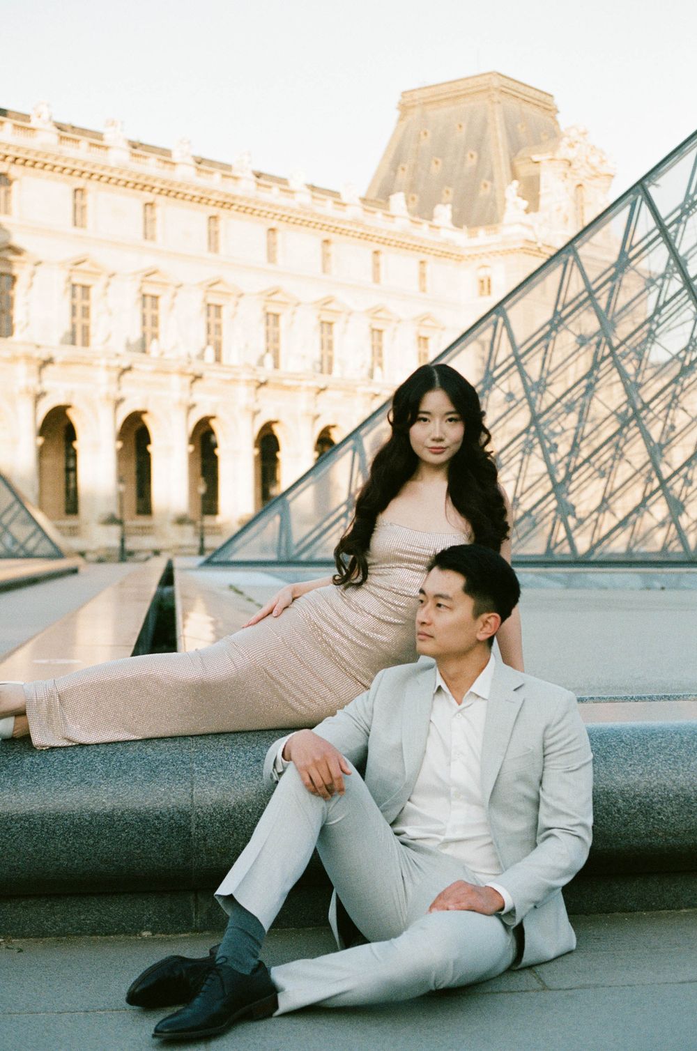 A couple poses elegantly near the glass pyramid at the Louvre Museum in Paris during sunset. editorial paris photographer.