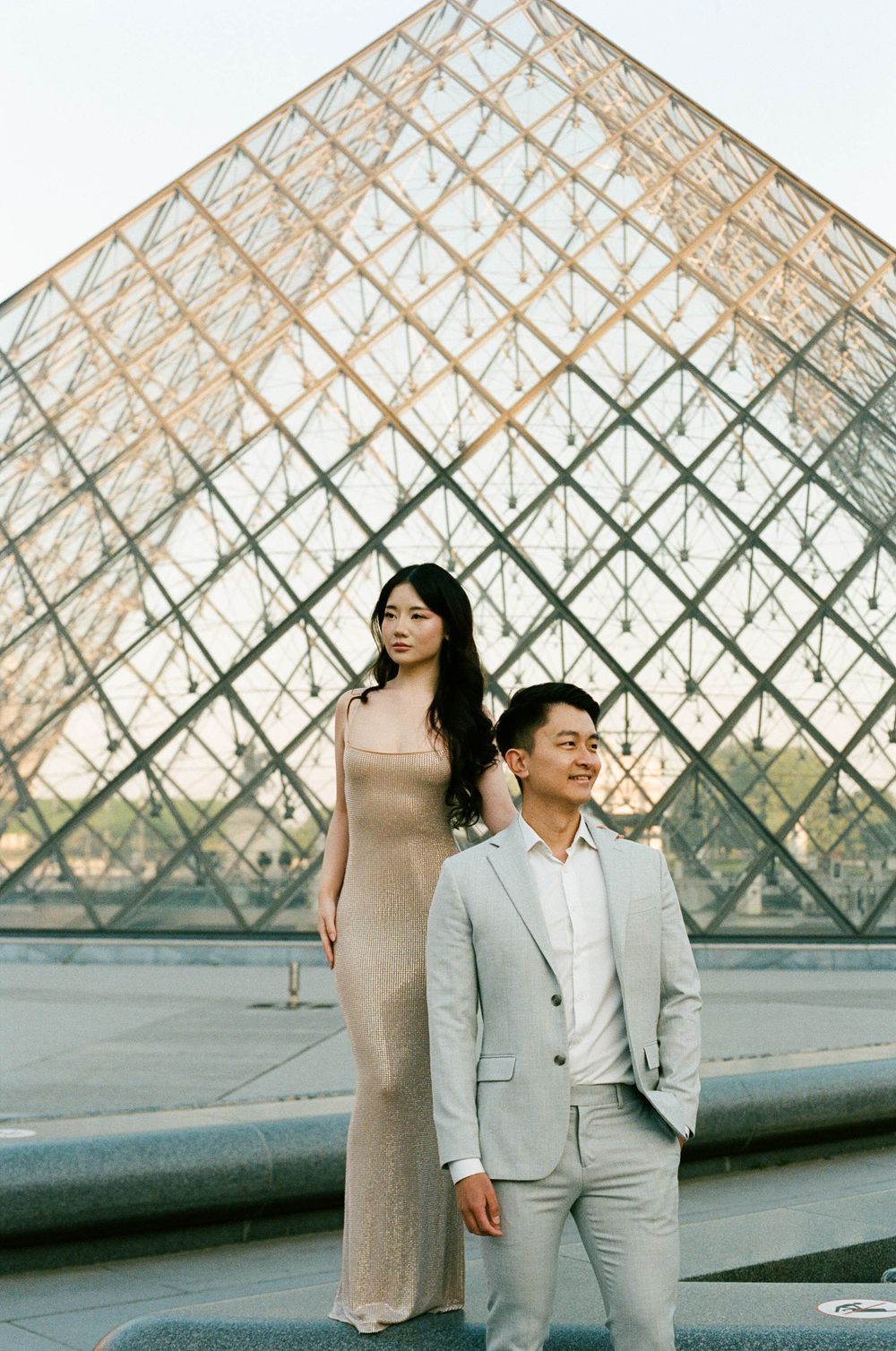 A couple poses elegantly in front of the glass pyramid at the Louvre Museum in Paris, France. paris film photographer. paris couples outfit