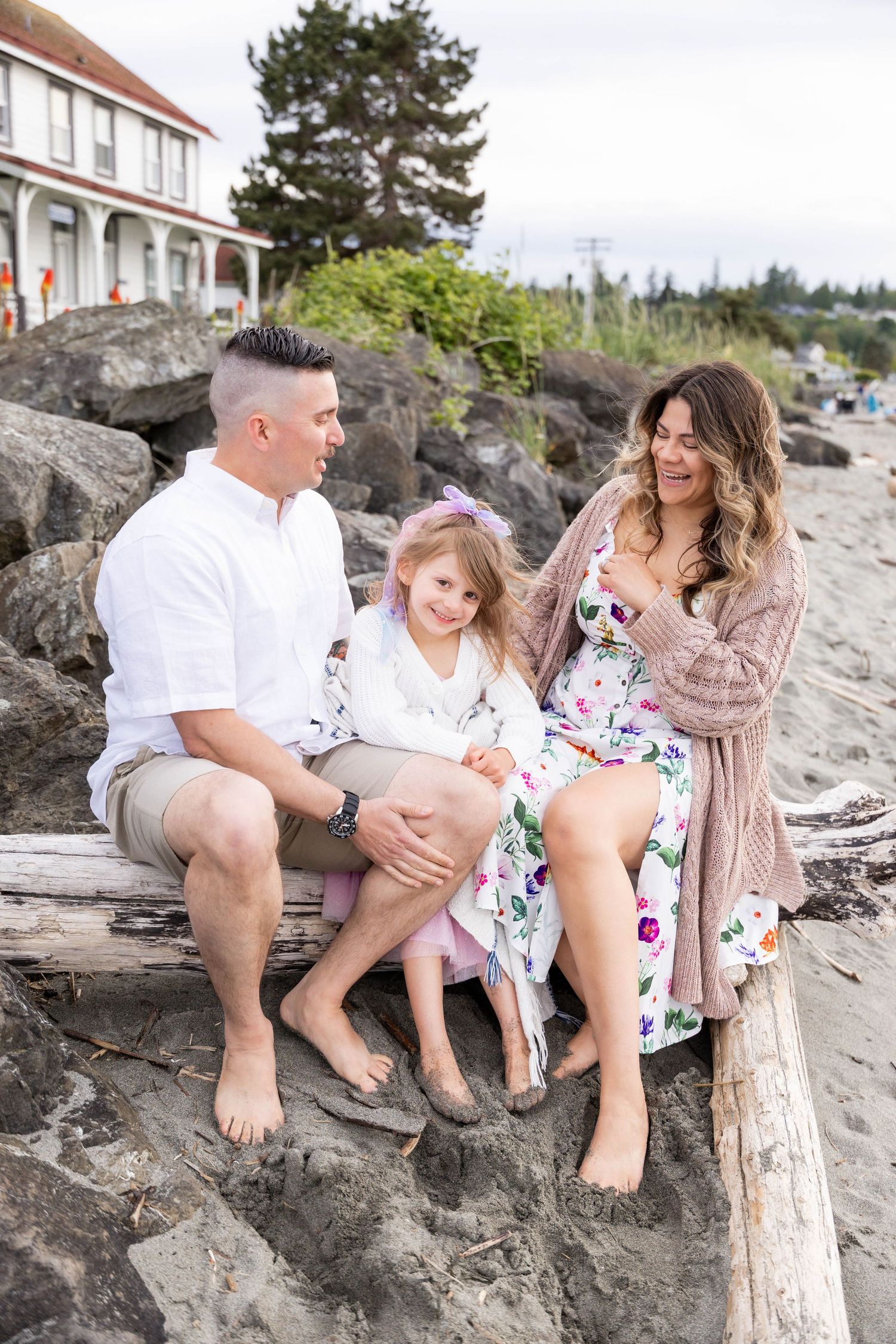 Three people sit together on rocky beach shore wearing casual summer outfits near a white house with trees in background.