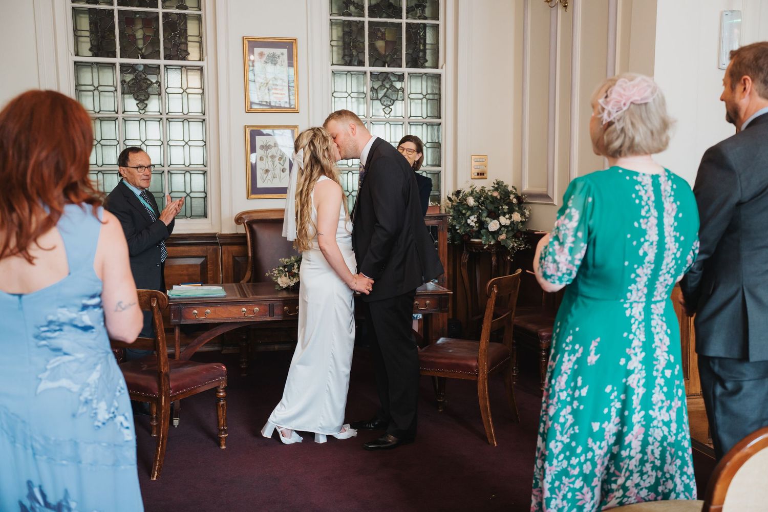 Bride and groom share their first kiss during an intimate registry office wedding ceremony with guests watching.