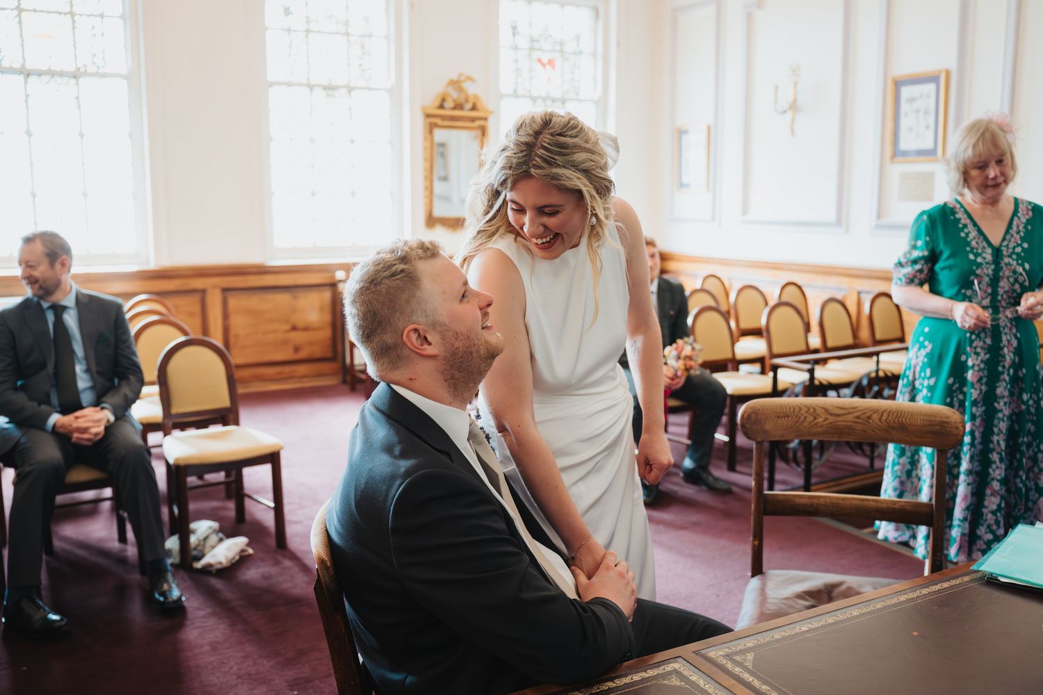 Bride in white gown laughs with seated groom during a relaxed civil wedding ceremony indoors at Oxford Registry Office