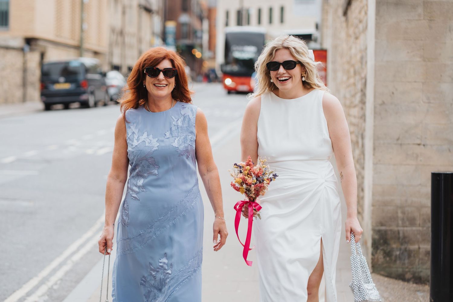 The bride is smiling and walking on a sunny Oxford city street, one in a wedding dress holding a colorful bouquet.