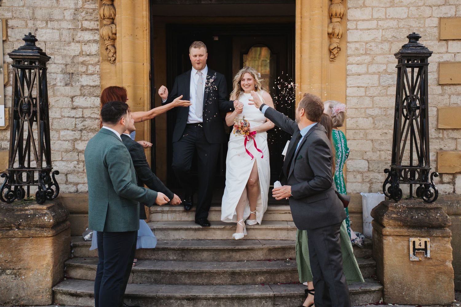 Bride and groom exit a grand stone building greeted by cheering wedding guests throwing confetti.