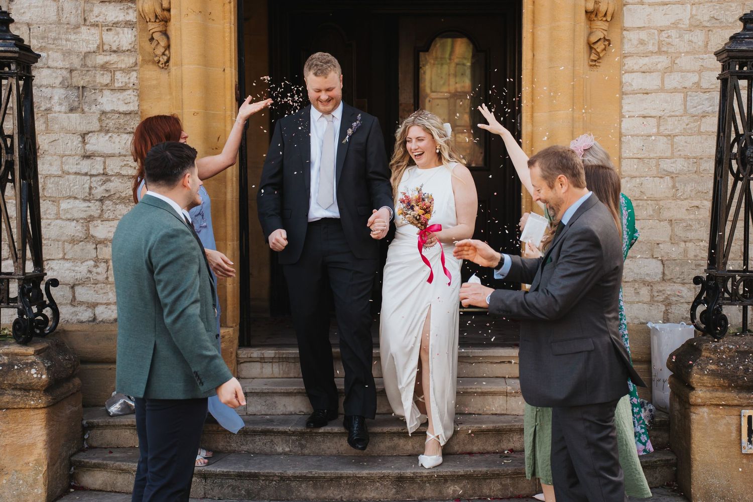 Smiling newlyweds descend stone steps as guests celebrate with confetti outside a classic stone venue.