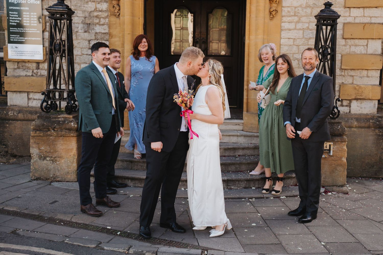 Newlywed couple share a romantic kiss outside Oxford Registry Office surrounded by smiling guests.