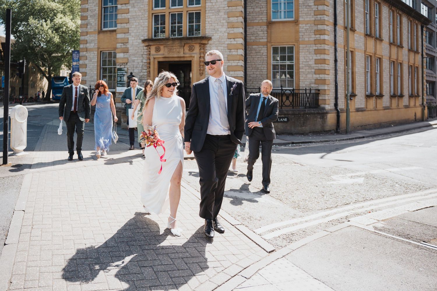 Bride and groom in sunglasses walk hand in hand through a sunny Oxford city street after their Oxford Registry Office wedding