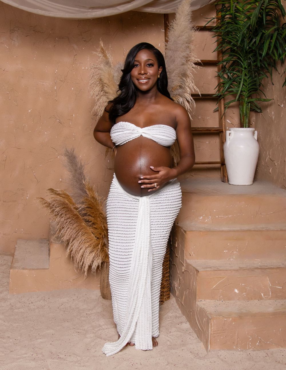 A figure poses in a white ruched off-shoulder dress against a textured brown wall with potted plants in the scene.