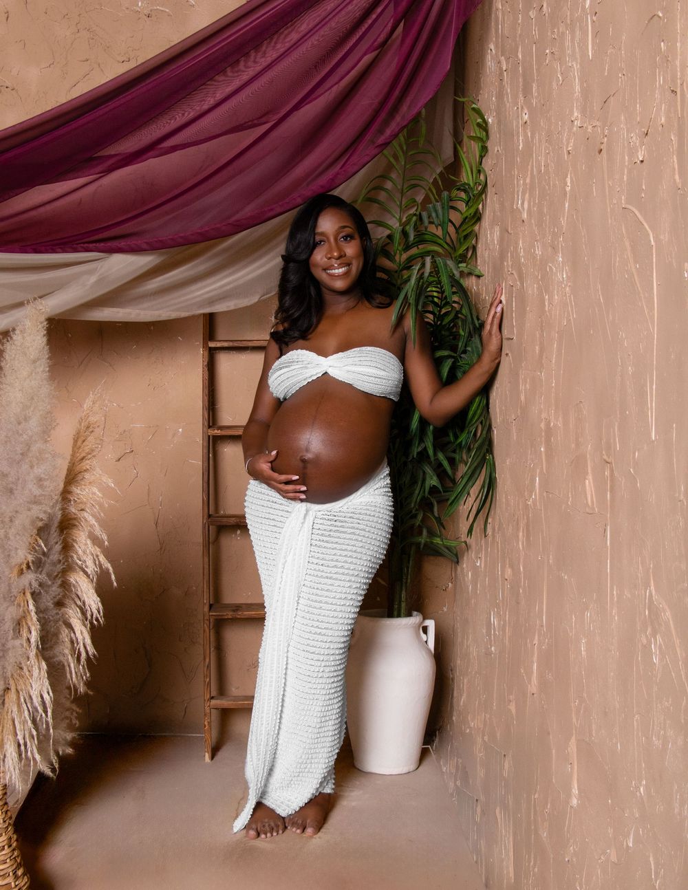 A maternity photo featuring an expectant parent in white attire posing against a beige backdrop with draped burgundy fabric.
