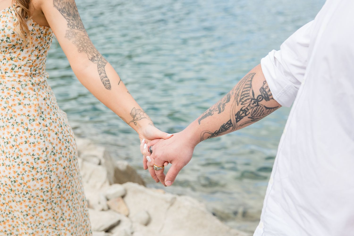 Close-up of tattooed couple holding hands by lake with rocky shoreline.