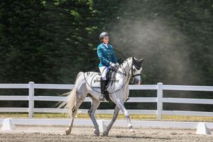 Equestrian rider in teal jacket on white horse performing dressage in outdoor arena with white fence.