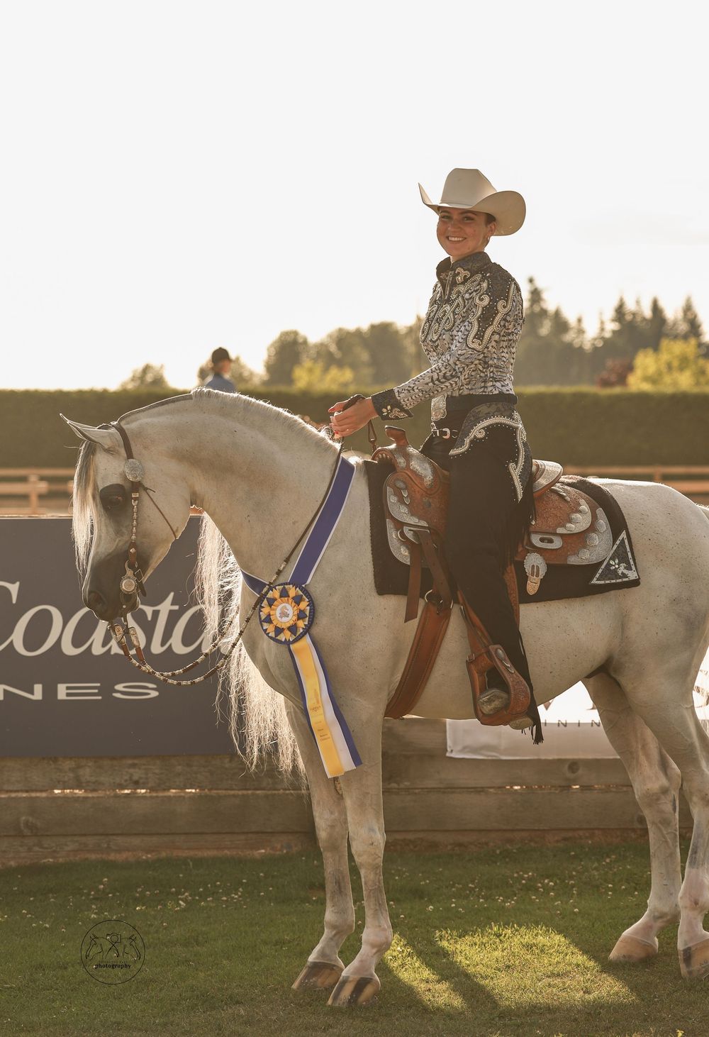 Smiling rider in cowboy hat on white horse displaying blue ribbon award at outdoor equestrian competition.
