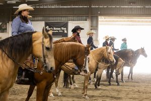 Riders on horseback practice western equestrian skills inside a dusty indoor arena wearing cowboy hats.
