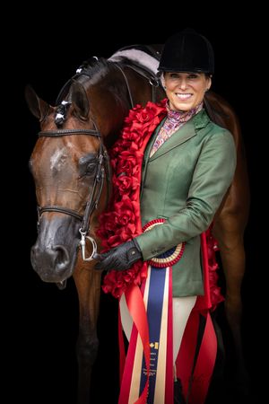 Smiling equestrian in green jacket holds award ribbons beside bay horse adorned with red rose garland on black background.