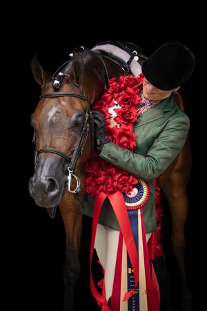Equestrian rider in green jacket hugging bay horse adorned with red roses and championship ribbon on black background.
