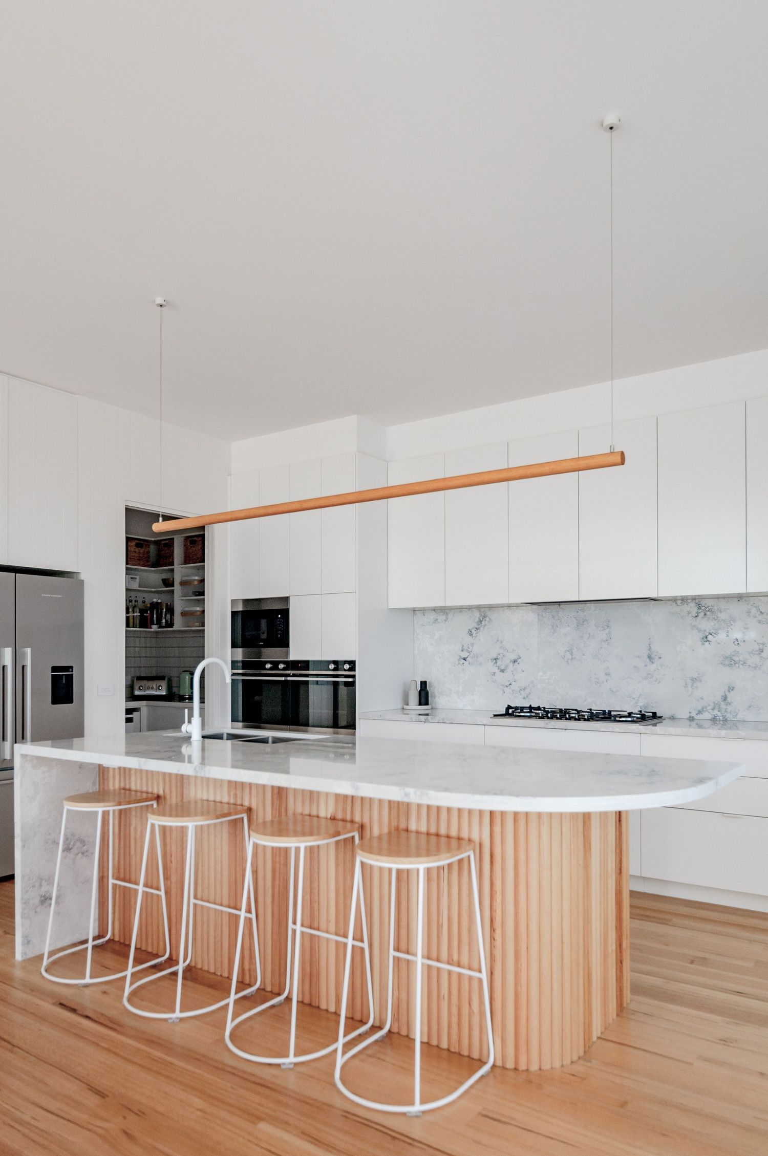 Modern white kitchen with marble countertops, wooden accents, and minimalist white bar stools at the island.