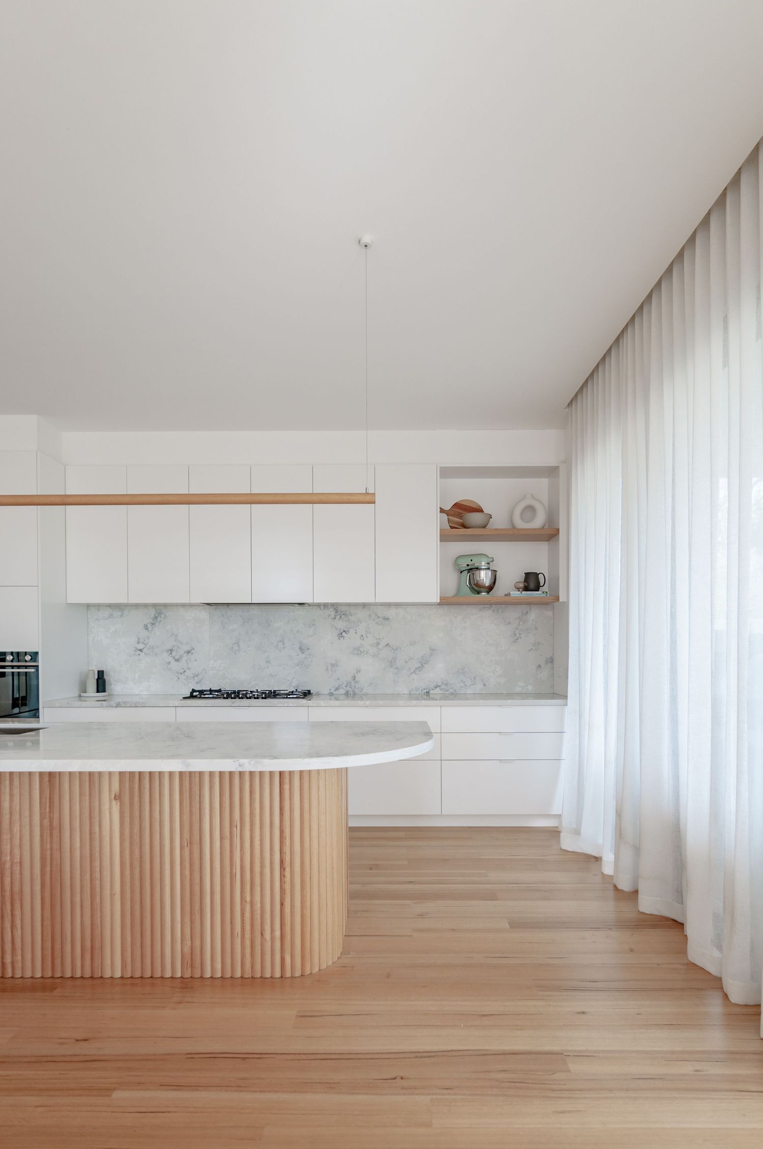 Modern white kitchen with curved wooden island, marble backsplash, and floor-to-ceiling sheer curtains.