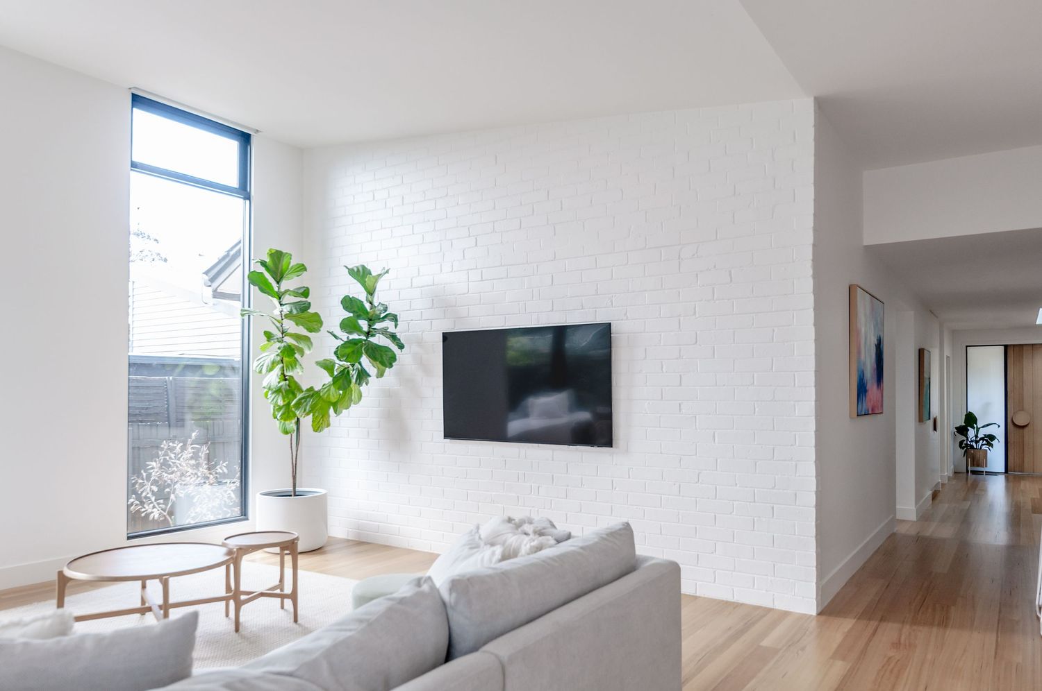 Minimalist living room with white walls, grey sofa, wall-mounted TV, and fiddle leaf fig plant by window.