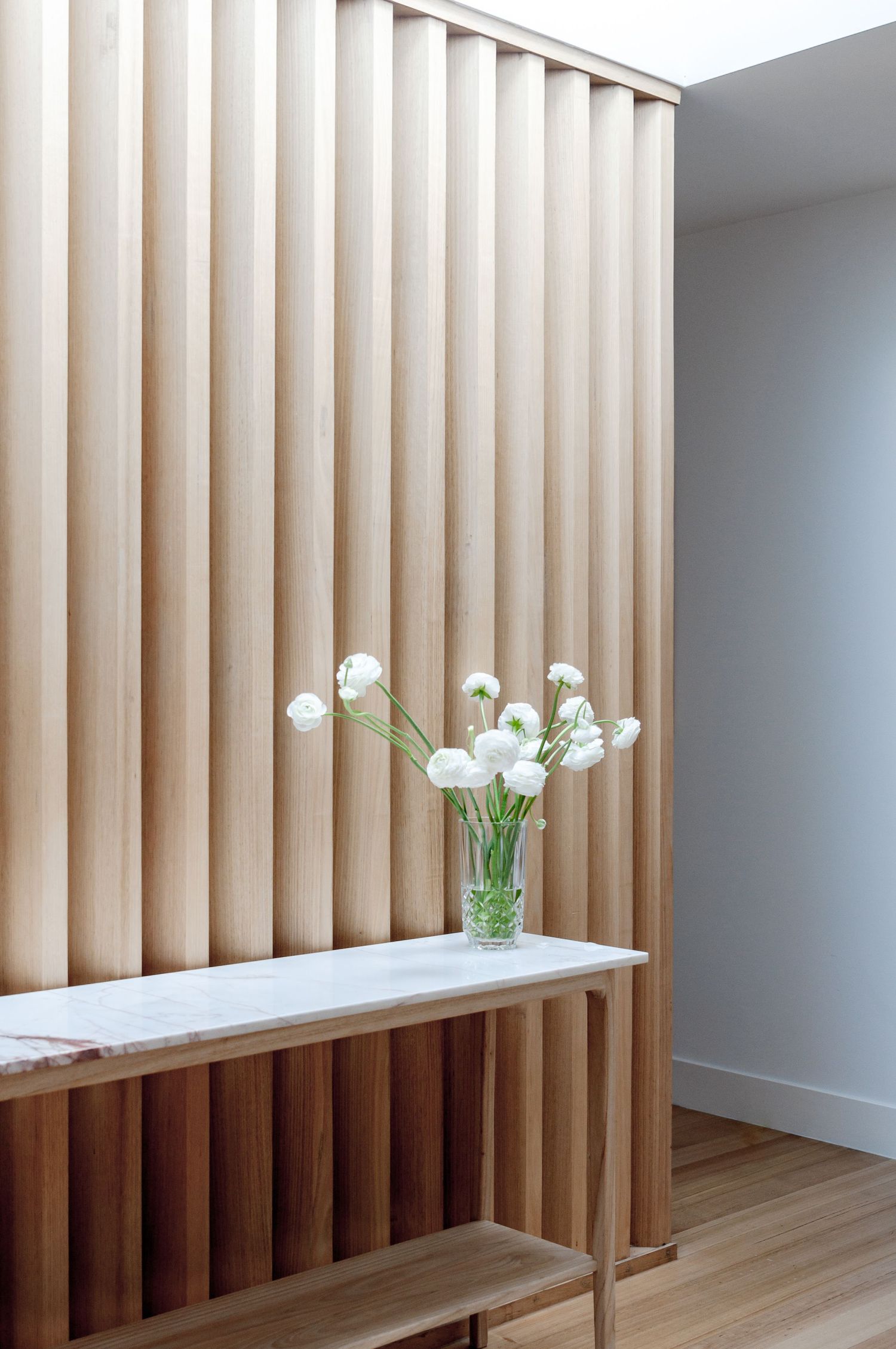 Elegant wooden slat wall feature with white console table and fresh flowers in glass vase.