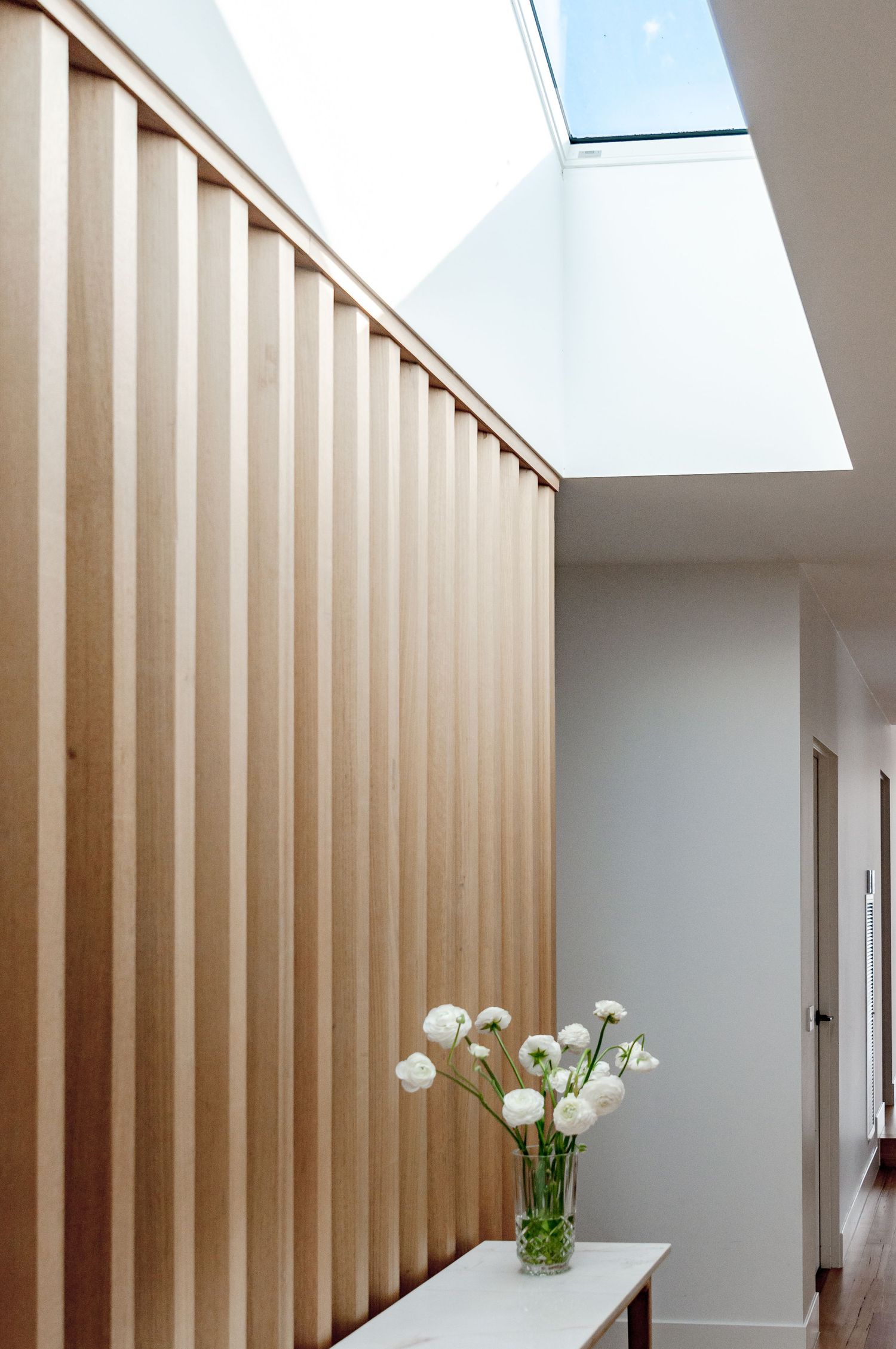 Architectural detail showing wooden slat wall meeting skylight with white console table and flowers below.