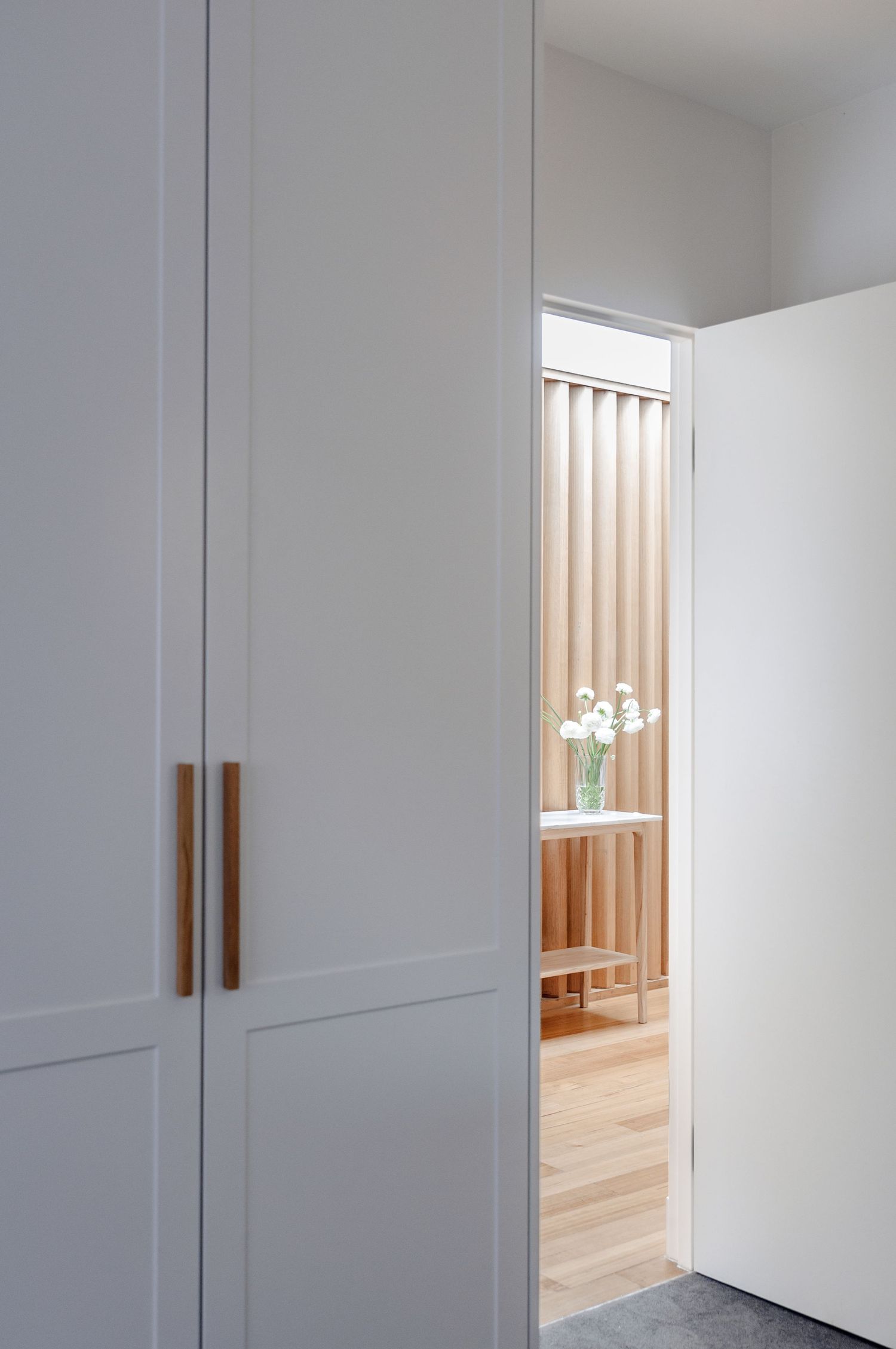 White built-in storage with wooden handles offering a glimpse of vertical slat feature through doorway.