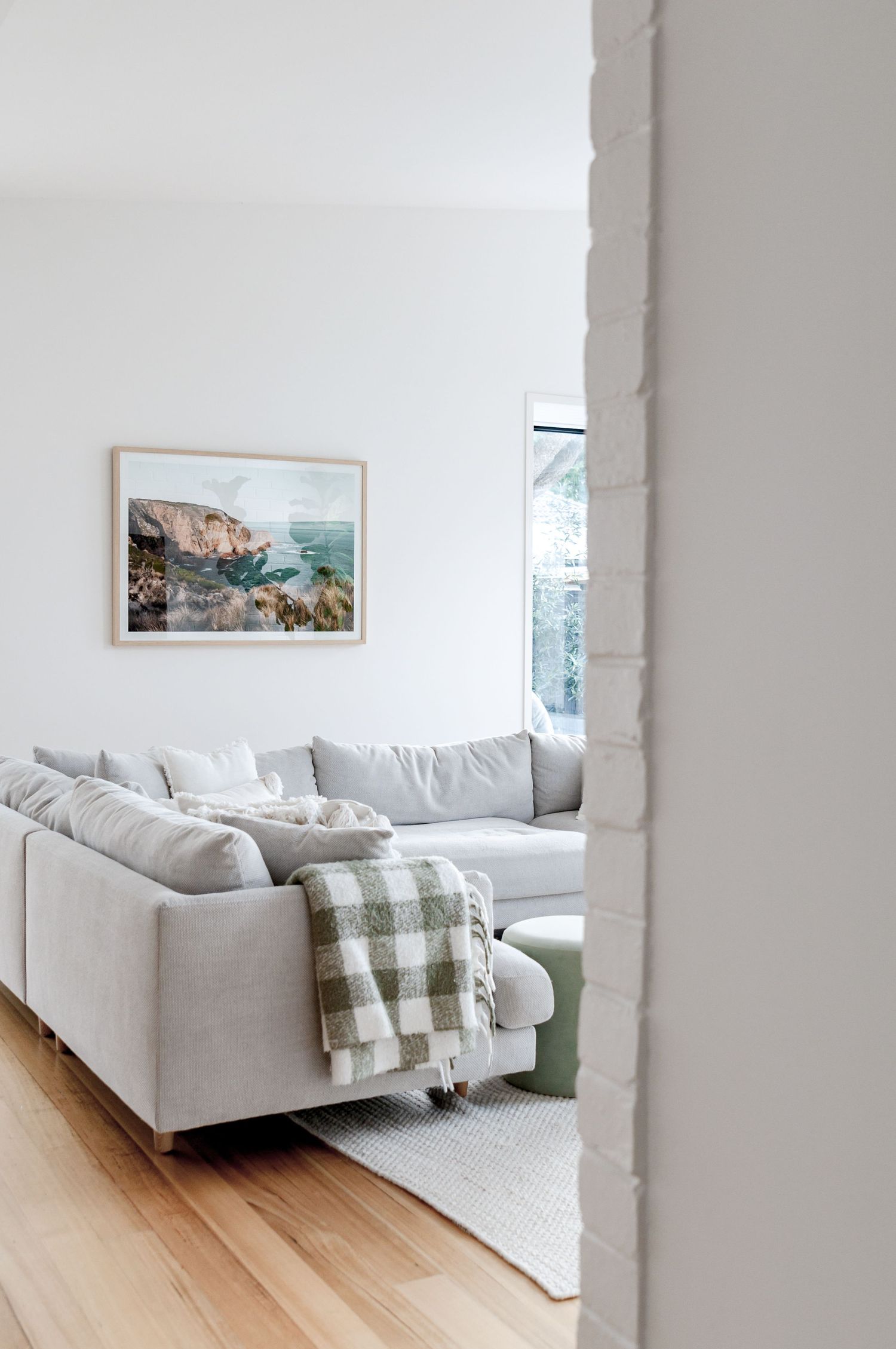 Cozy living room corner with light gray sectional sofa, plaid throw, and coastal artwork on white walls.