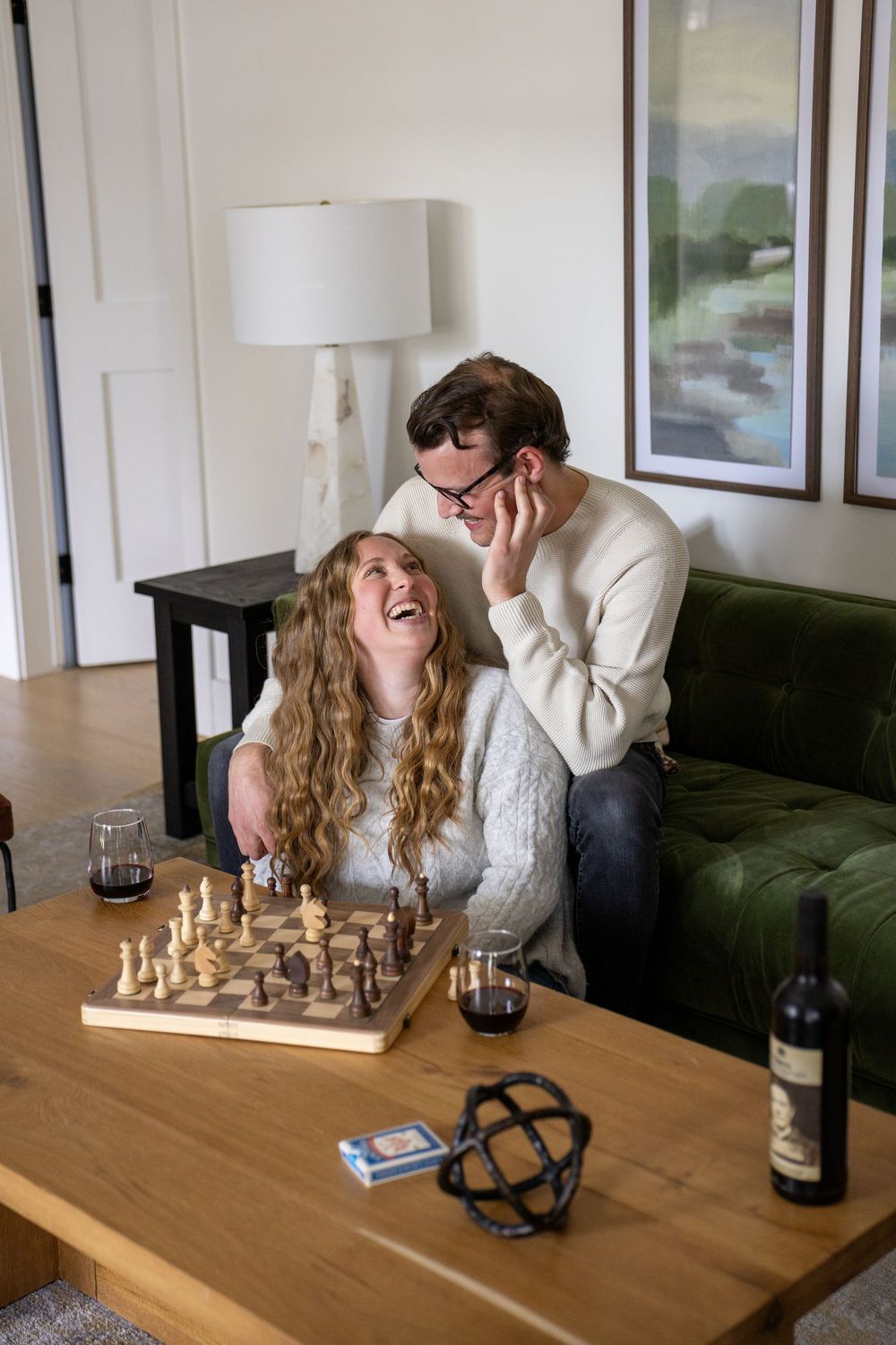 Two people laugh together while playing chess at a wooden table with wine glasses and decor.