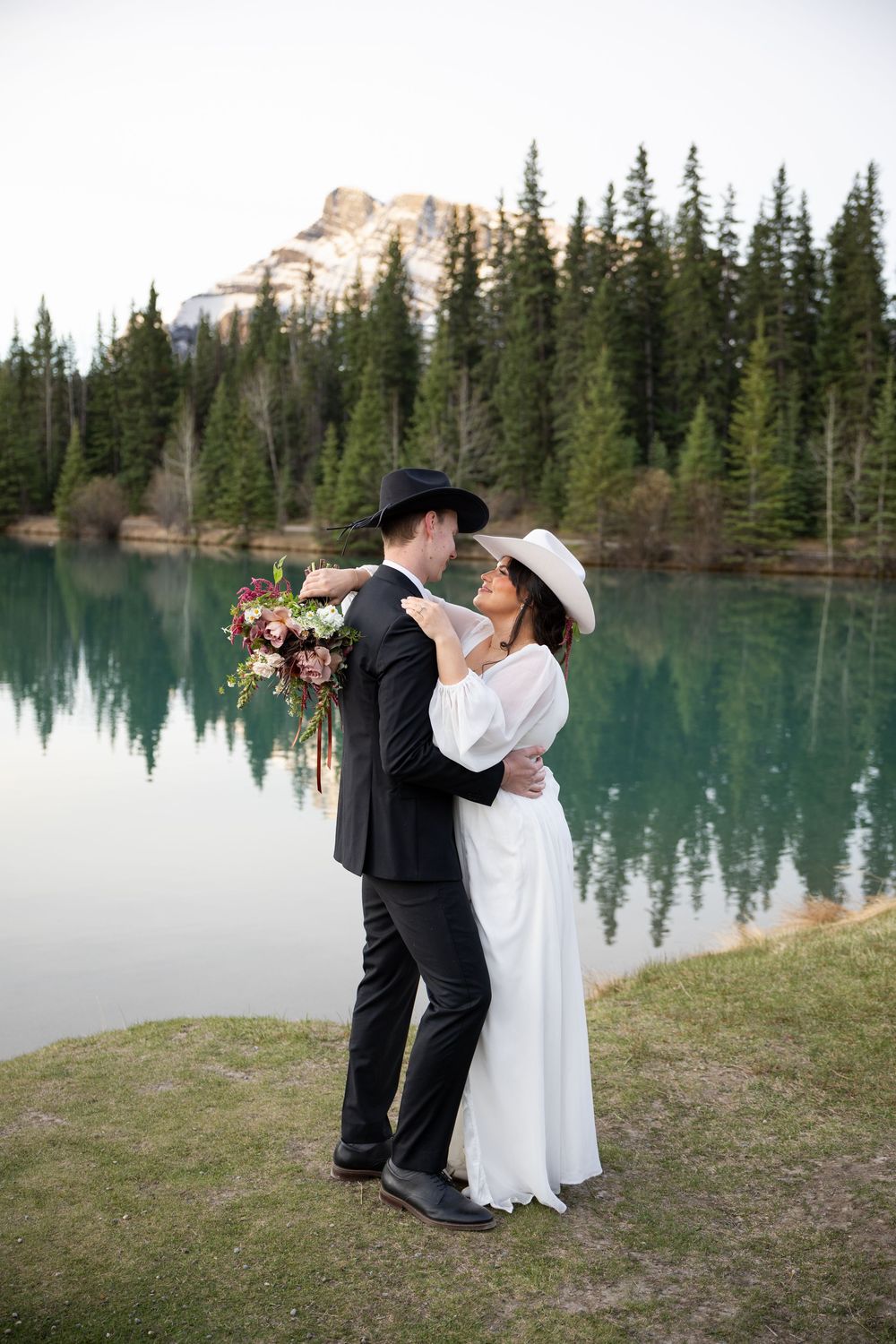 A couple in wedding attire shares a romantic moment beside a turquoise mountain lake surrounded by evergreen trees.