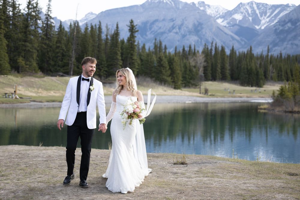 A couple walks along a scenic mountain lake with snow-capped peaks and evergreen trees in the background.