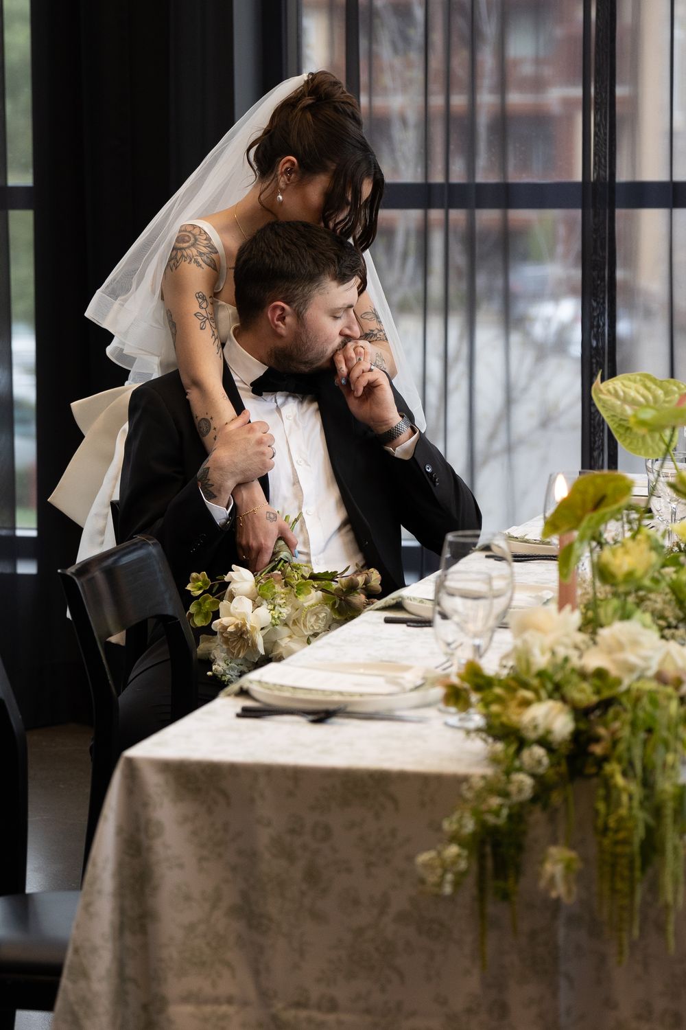 A couple shares an intimate moment at their wedding reception table decorated with greenery and white linens.