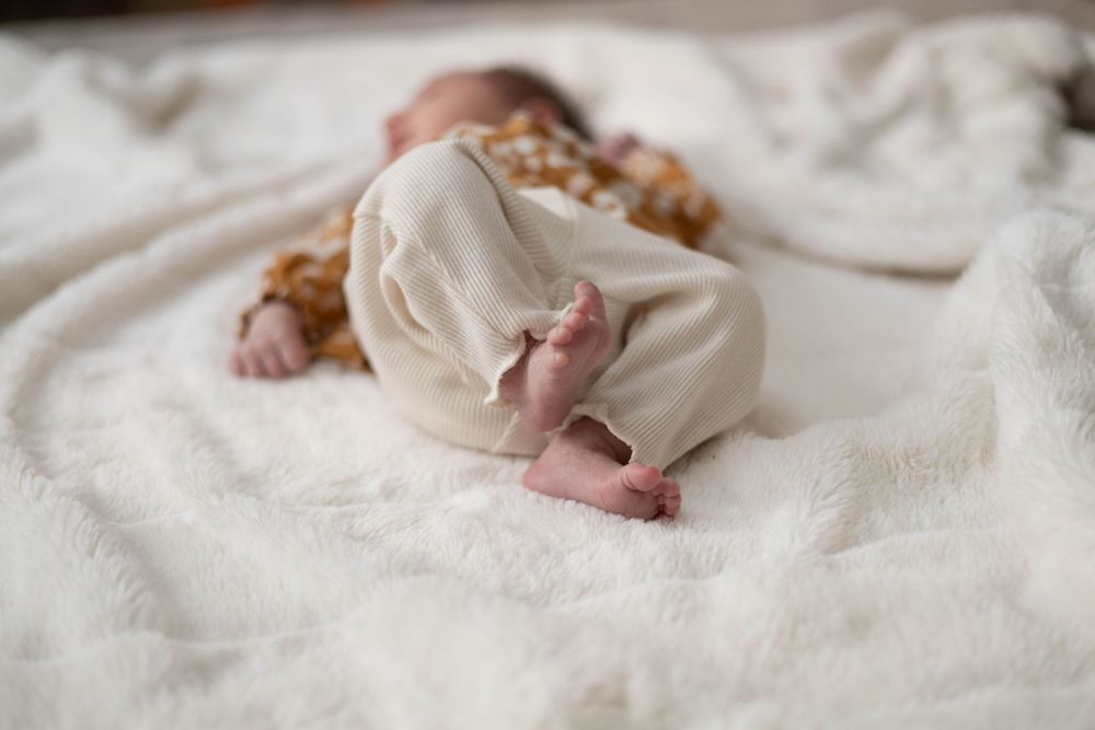 Small baby feet peek out from underneath a cream colored blanket on white bedding.