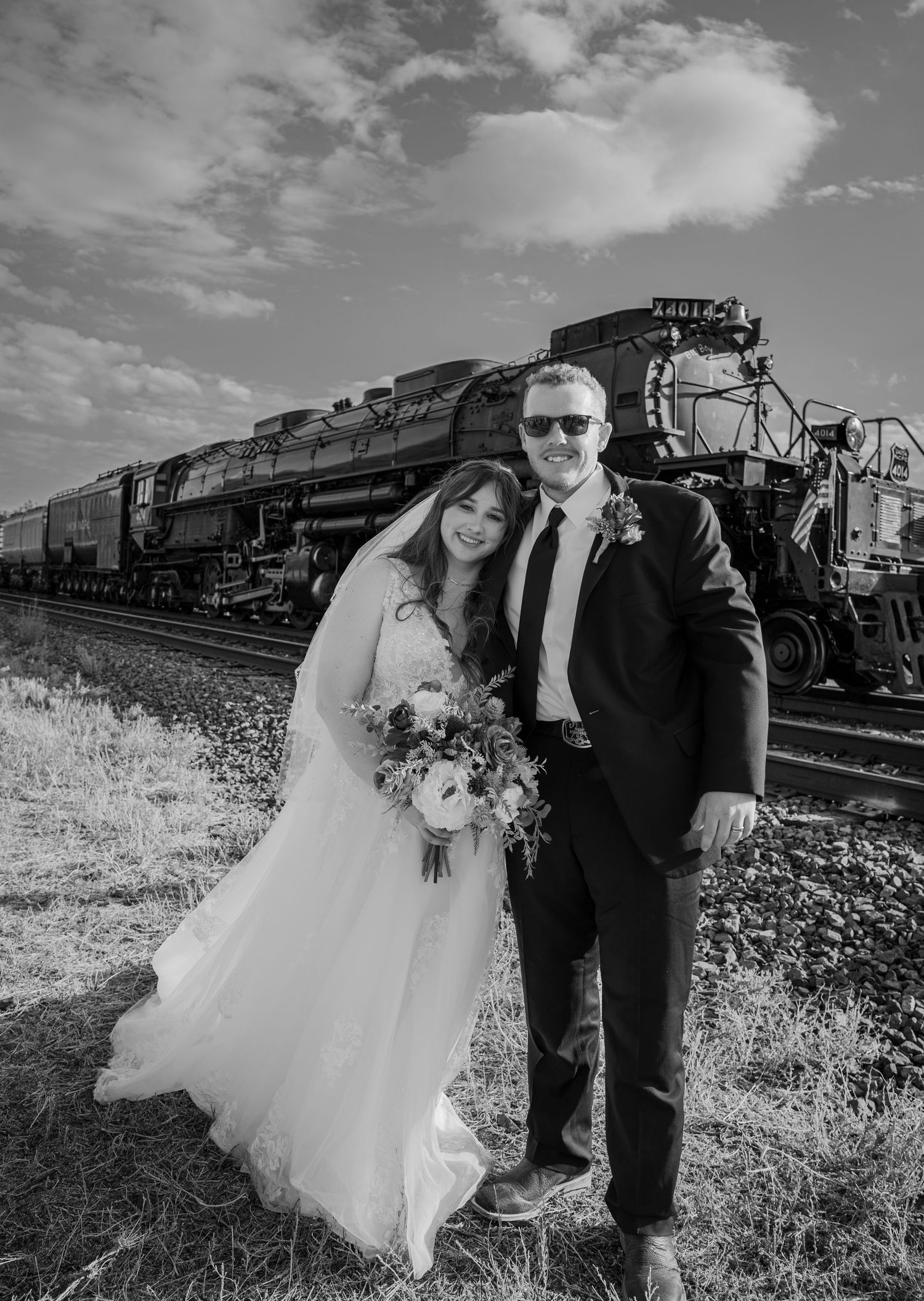 A wedding couple poses in front of a vintage steam locomotive in black and white.