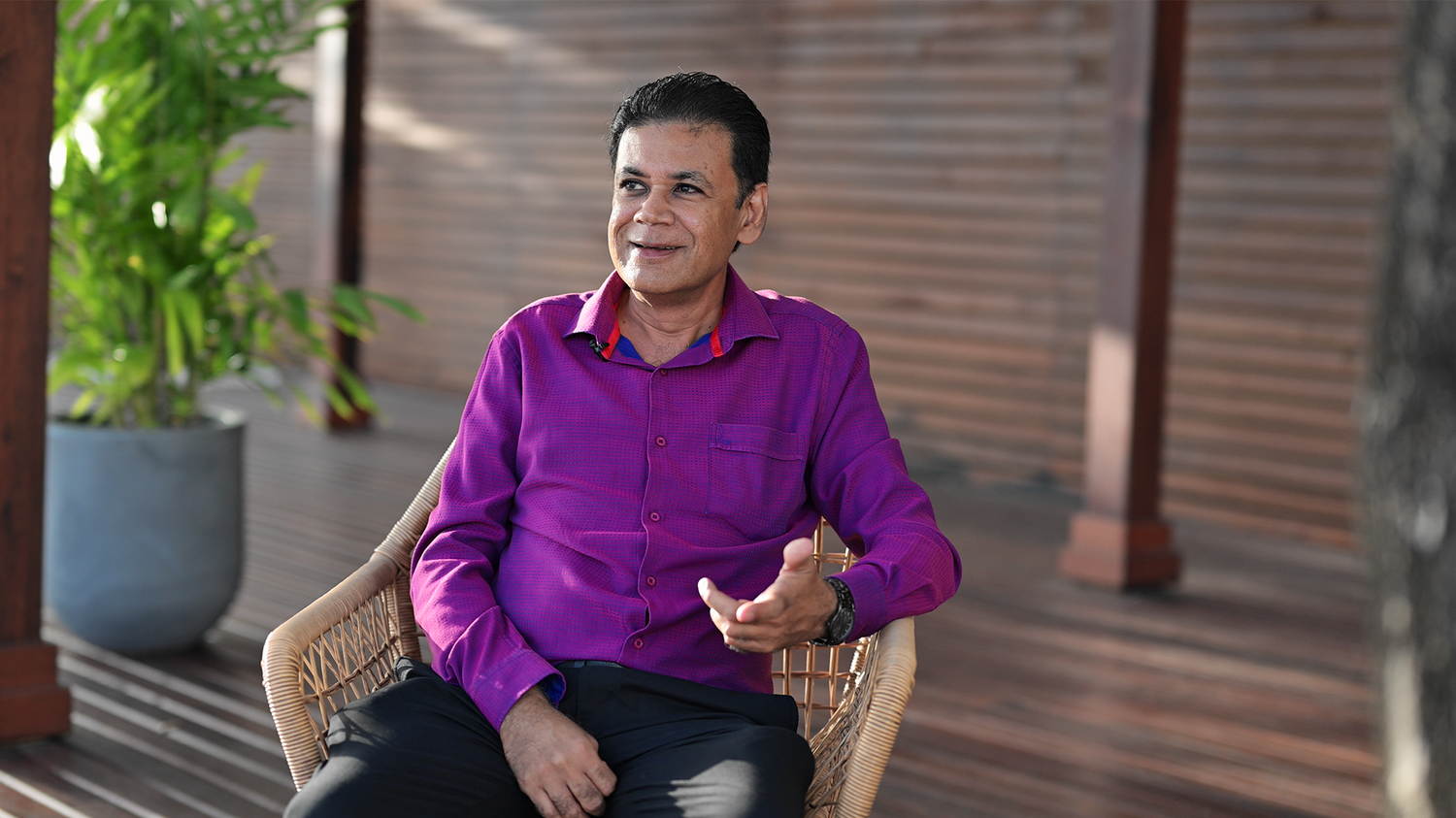 Person in purple polo shirt seated in wicker chair smiling against outdoor setting with plants.