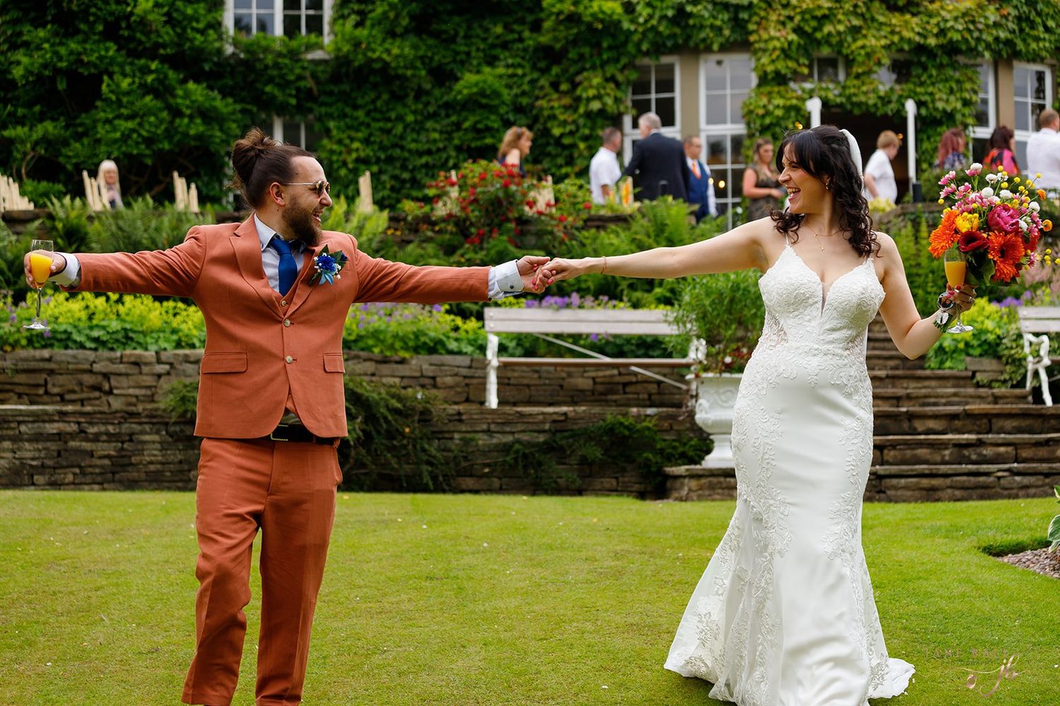 Bride in white gown and groom in orange suit hold hands outdoors at a garden wedding venue. Hilltop Country House.