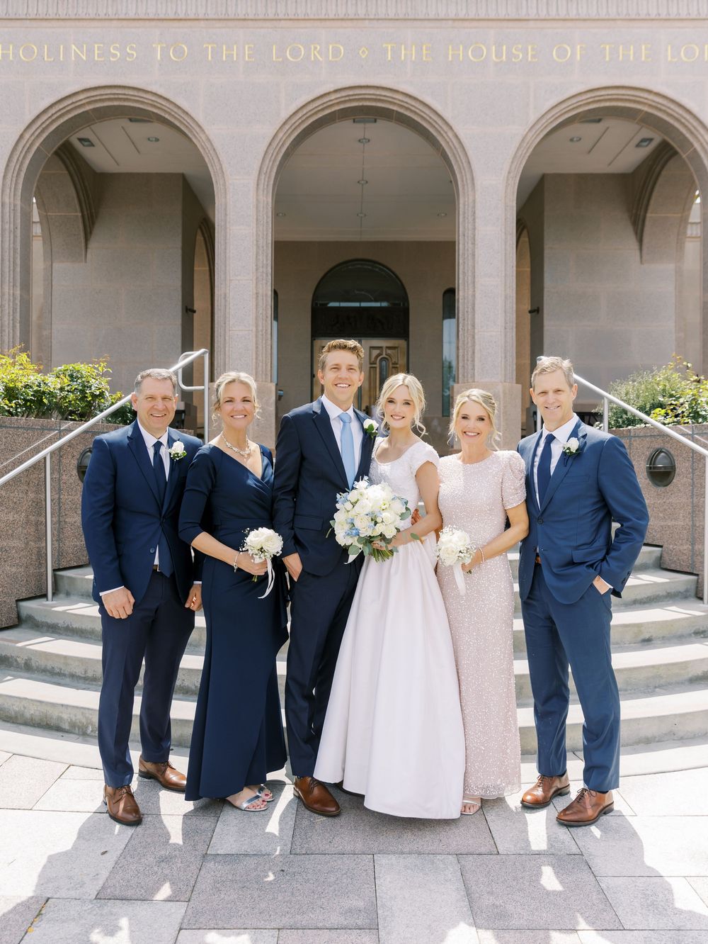 Wedding party in navy suits and light dresses posing under elegant stone archways.