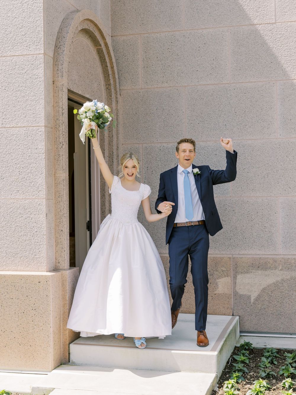 Wedding couple and wedding party celebrate joyfully in front of stone archway.