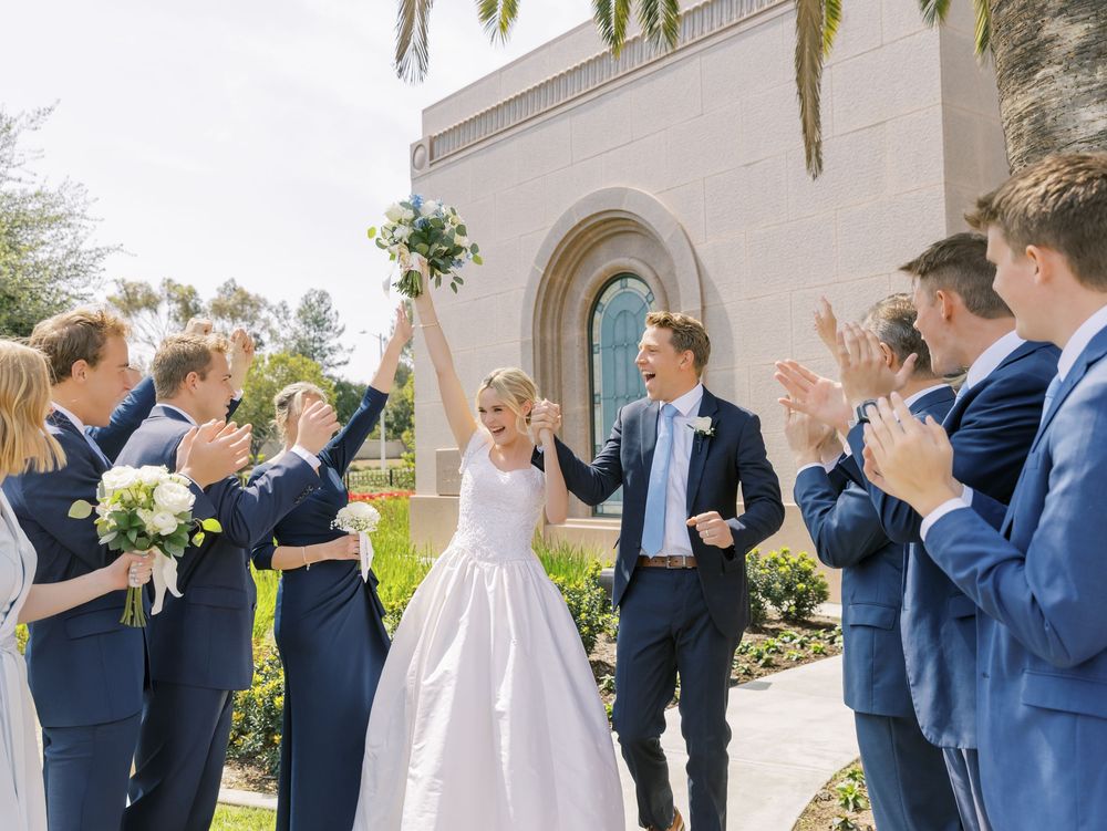 Wedding party in navy suits and light dresses celebrating with raised arms outdoors.
