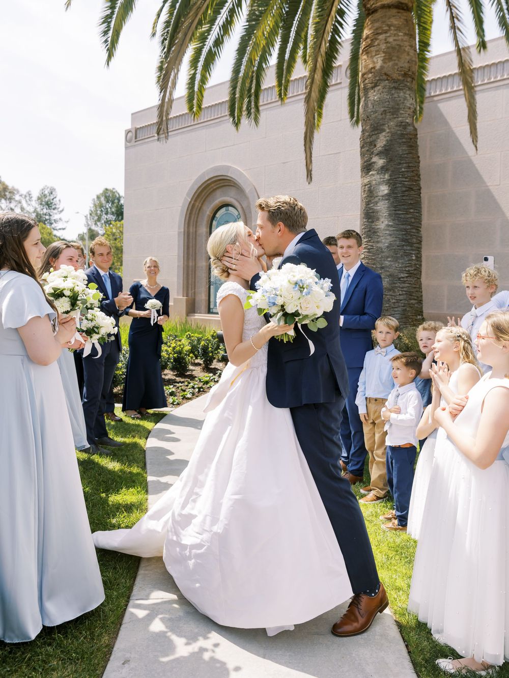 Wedding party in white dresses and navy suits poses on church lawn with palm trees.