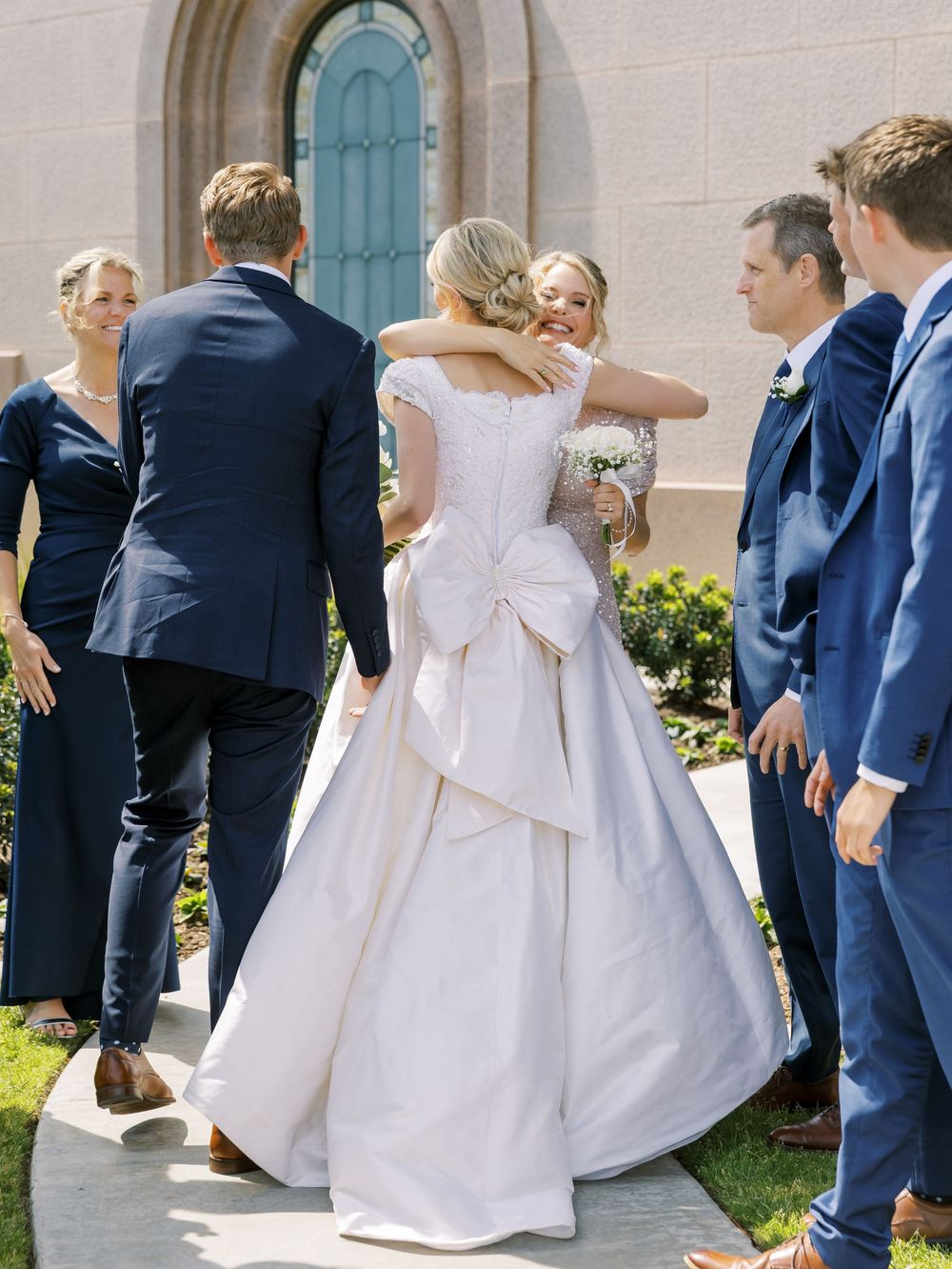 Wedding party members in navy suits and white dresses walk together in a playful line outside a building.