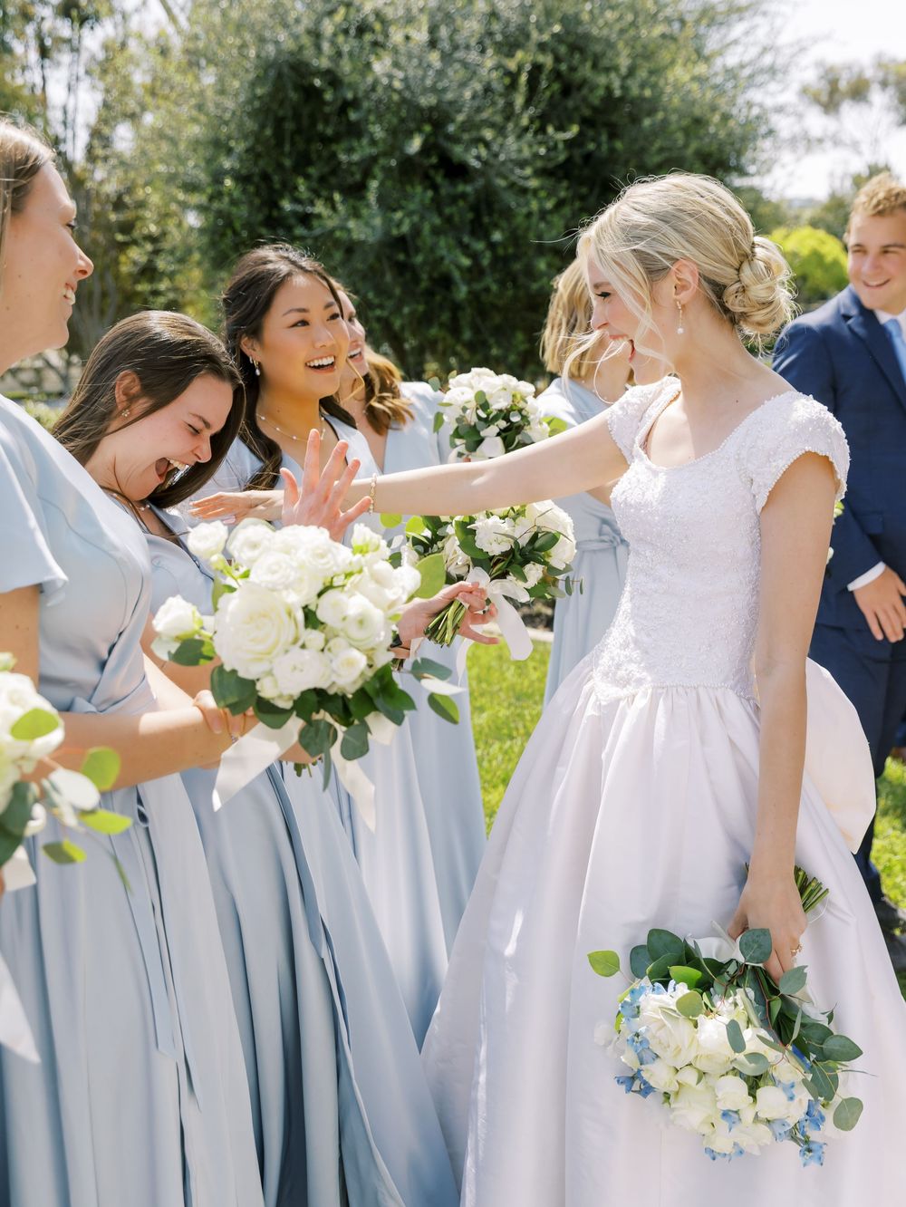 Bridesmaids in powder blue dresses laughing together while holding white floral bouquets.