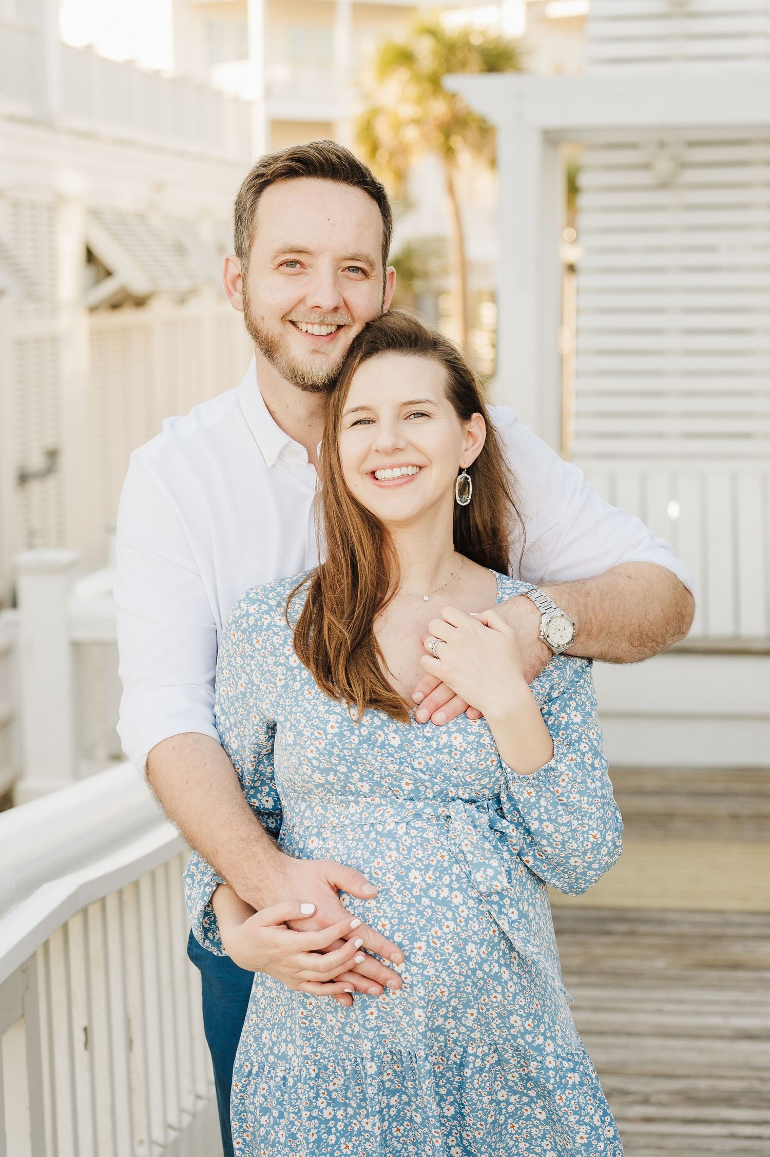 A couple in coordinated blue and white outfits embrace lovingly on a white porch during an outdoor photo session.