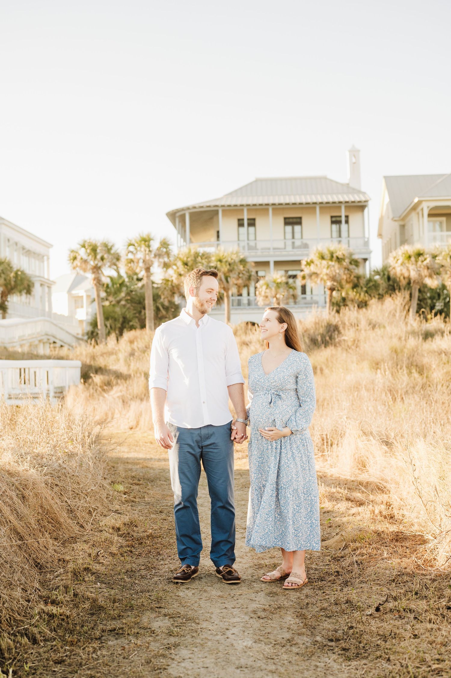 Couple standing together in golden beach grass at sunset with coastal homes in background.