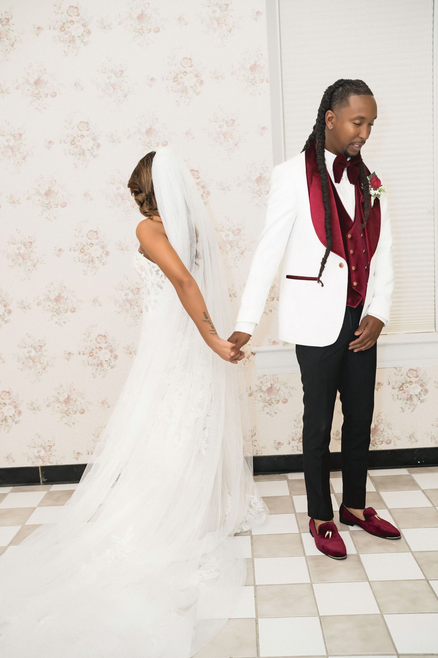 Two people in formal attire stand on a tiled floor, one wearing a white dress and one in a burgundy vest.