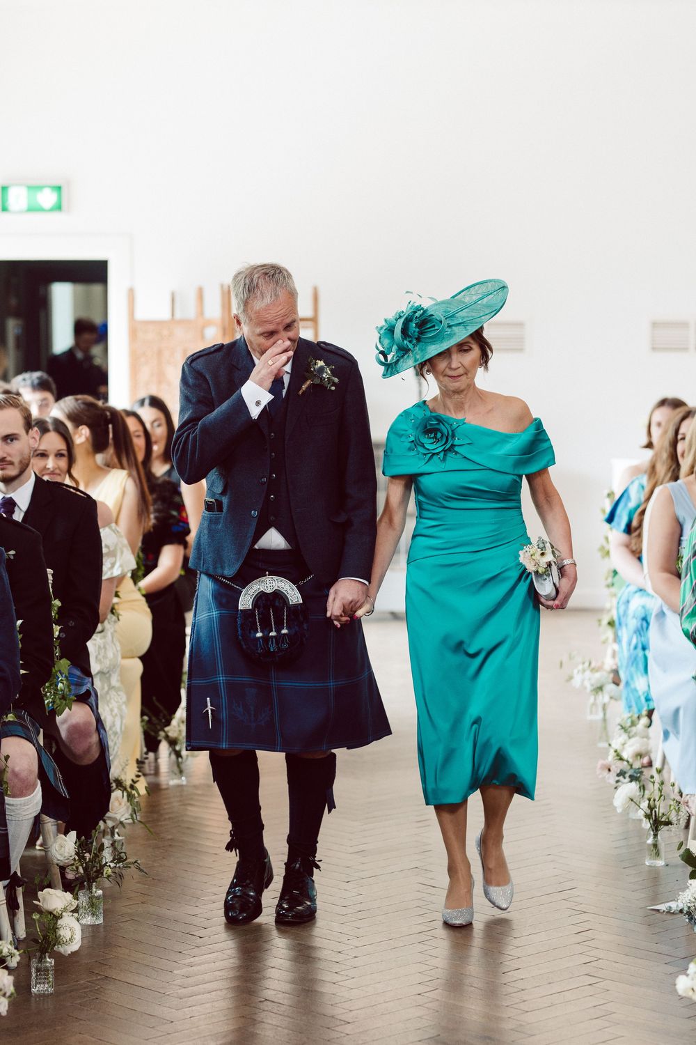 Wedding party walks down aisle in coordinated outfits of black kilts and a striking turquoise dress.