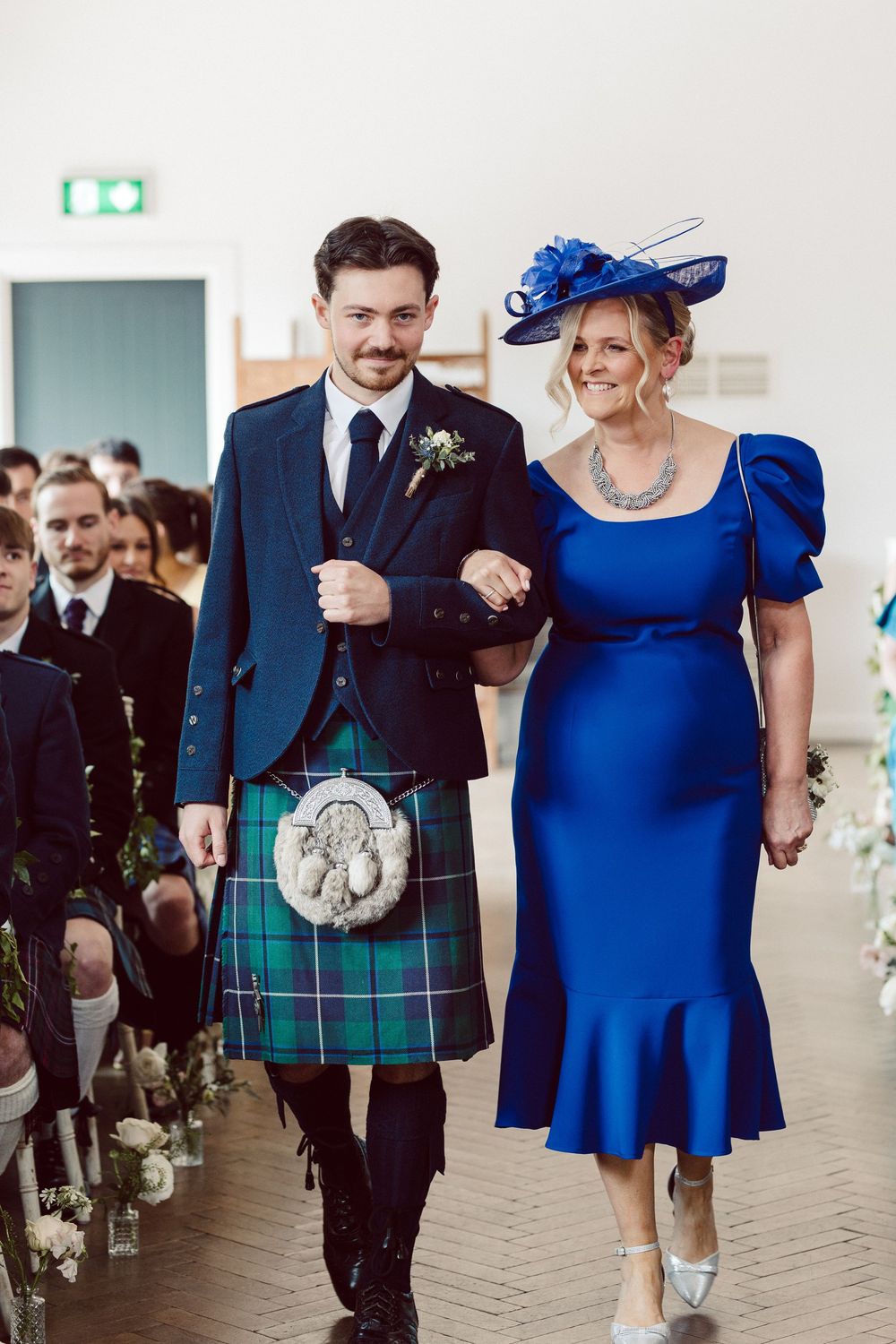 Wedding party members in formal attire walk down the aisle with men wearing kilts and women in royal blue dresses.