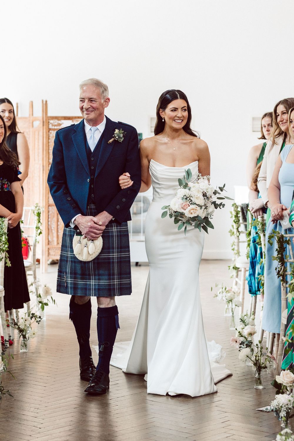 Wedding processional with bridesmaids in blue dresses and groomsmen in kilts walking down aisle decorated with flowers.