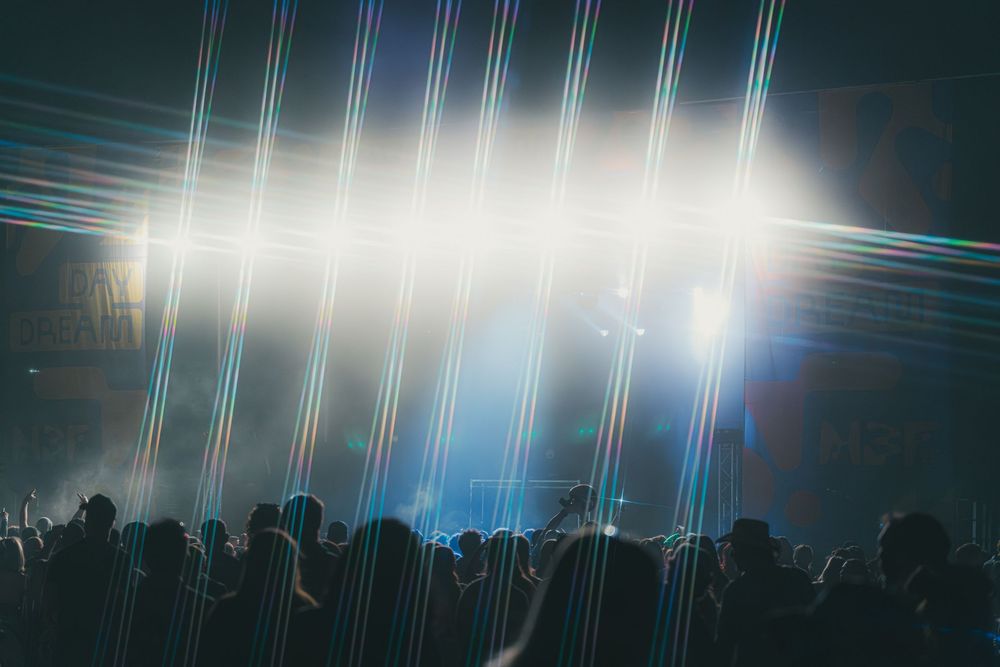 A silhouetted crowd stands beneath bright stage lights creating dramatic vertical light beams at a concert venue.