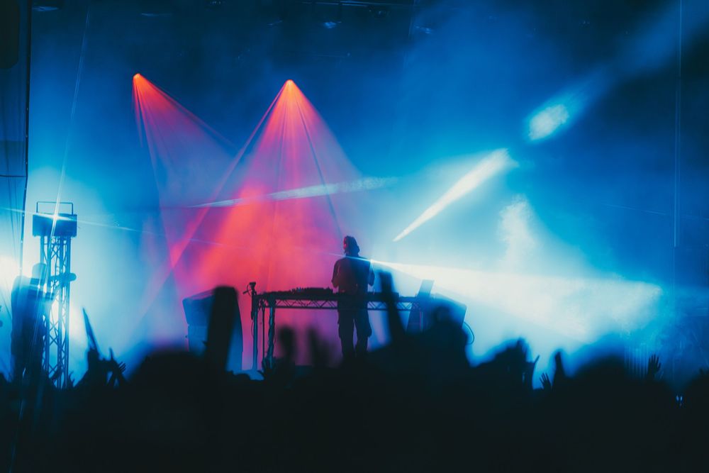 Colorful concert stage lights illuminate a dark silhouetted crowd with blue and pink hues creating a vibrant atmosphere.