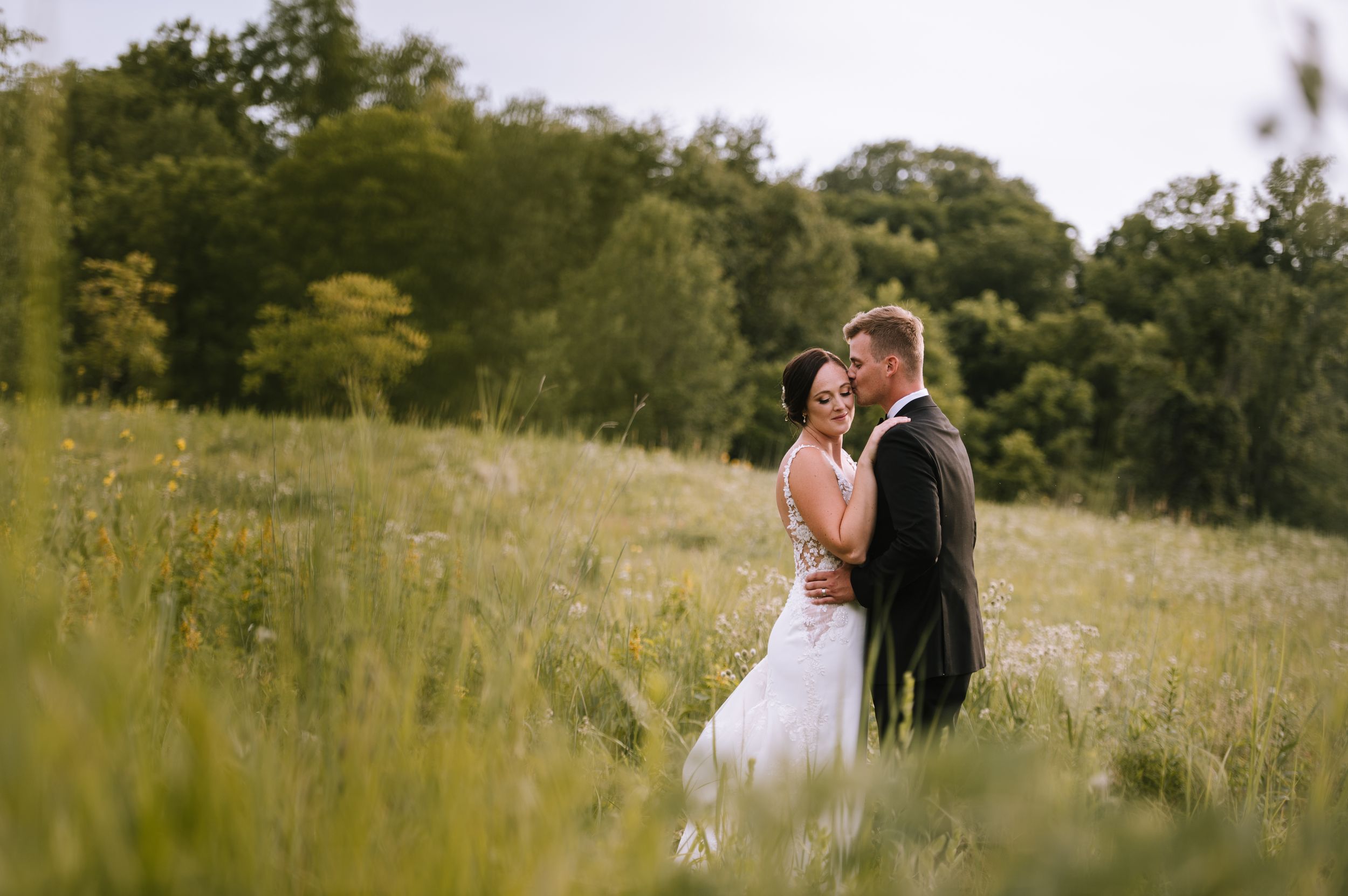 A couple in wedding attire stands together in a sunlit meadow with tall grass and trees in the background.